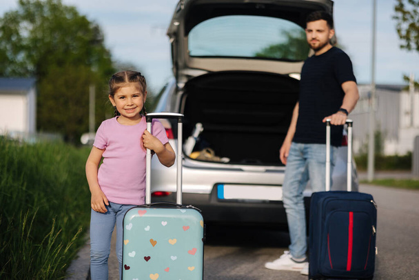 Papá y su hija están de pie con sus maletas cerca de un coche con un baúl abierto. Niña feliz preparándose para viajar. Foto de alta calidad - Foto, Imagen