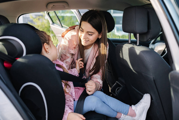 Attractive young mother put her daughter in a car seat and fastens her seat belts. Woman care about kid. Protection during the trip in the car. High quality photo - Photo, image
