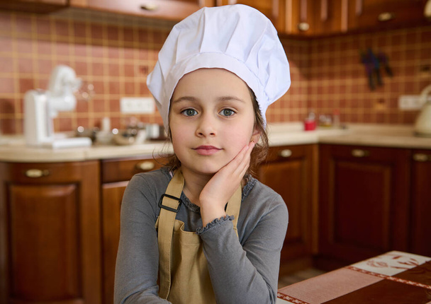 Authentic portrait of Caucasian adorable child girl in white chef hat and beige apron, little baker confectioner looks at camera, standing at home kitchen interior. Cooking class. Kids learn culinary - 写真・画像