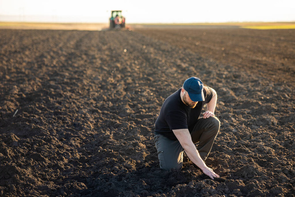 Jeune agriculteur examinant la saleté pendant que le tracteur laboure le champ
 - Photo, image