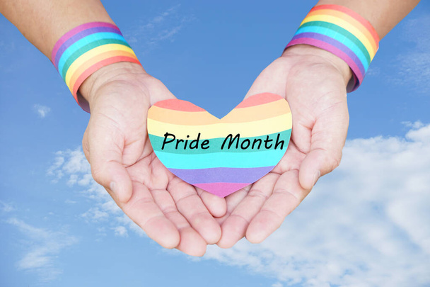 Close up hands wears rainbow color wrist band and holds rainbow heart shape with word Pride Month. Blue sky background. Concept, Pride month for LGBTQ community celebration in June. Human right   - Photo, Image