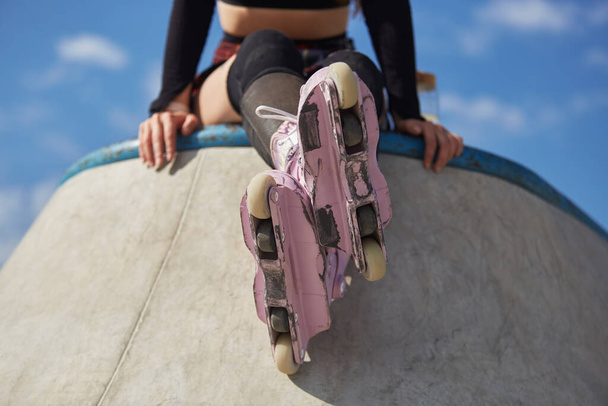 Young roller blader girl sitting on a concrete ramp wearing modern aggressive inline skates on her feet. Unrecognizable female athlete relaxing after a ride in a skatepark - Fotó, kép