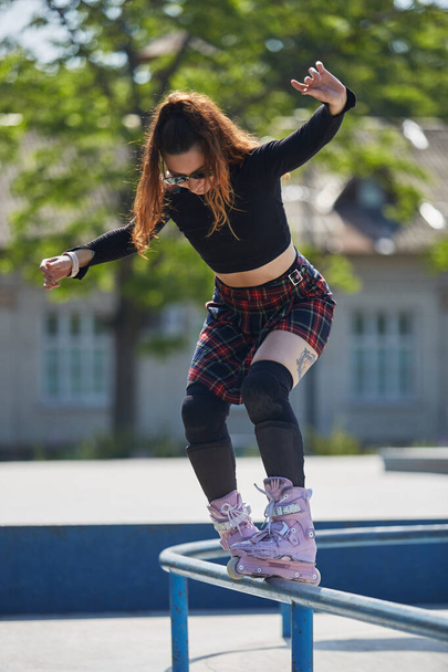 Cool young female grinding on a rail with aggressive inline skates. Tattooed chick skating in a skatepark outdoor in a sunny summer day. Roller blader performing soul grind trick on a handrail - Foto, imagen