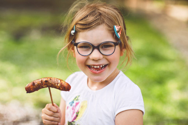 A kis óvodás lány grillezett kolbászt eszik. Boldog gyerek barbecue-n vagy picknicken. Egészséges ételek, családi nyaralás - Fotó, kép