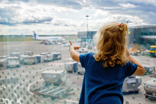 Cute little toddler girl at the airport, traveling. Happy healthy child waiting near window and watching airplanes. Family going on summer vacations by plane - Photo, Image