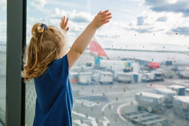 Cute little toddler girl at the airport, traveling. Happy healthy child waiting near window and watching airplanes. Family going on summer vacations by plane - Foto, immagini