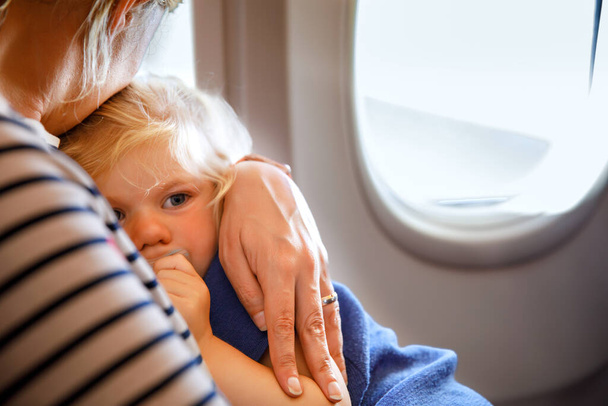 Жінка подорожує з маленькою дитиною на літаку. Сумна втомлена дівчинка, яка сидить з мамою біля вікна літака. Концепція материнства. Плаче дитина
 - Фото, зображення