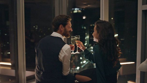 Attractive bearded man flirting with smiling woman drinking champagne late evening indoors. Two lovers relaxing together enjoying romance. Happy couple have pleasant talk looking on night city view. - Photo, Image
