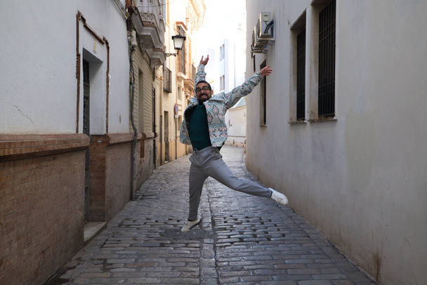A handsome young man with a beard and glasses is jumping for joy in a narrow street. Happiness and joy concept. - Foto, imagen