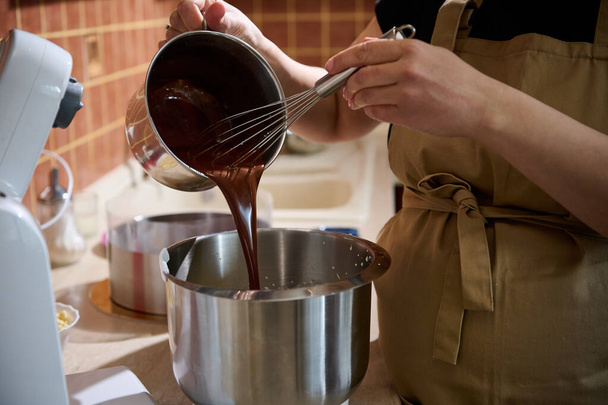 Close-up pastry chef pouring melted chocolate into a metal bowl of planetary mixer, mixing it with cream for preparing delicious chocolate cream or with whipped egg whites for making sponge biscuit - Φωτογραφία, εικόνα