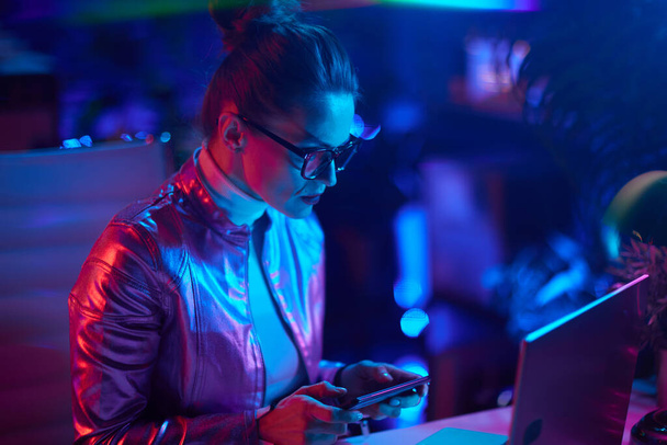 Neon metaverse futuristic concept. elegant female in glasses with laptop using smartphone in modern office. - Фото, зображення