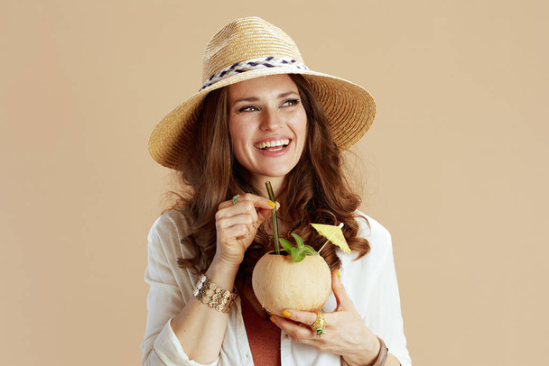 Beach vacation. smiling modern housewife in white blouse and shorts isolated on beige background with coconut cocktail and summer hat. - Foto, imagen