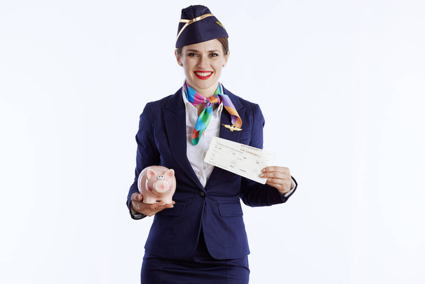happy modern female air hostess isolated on white background in uniform with flight tickets and piggy bank. - Foto, immagini