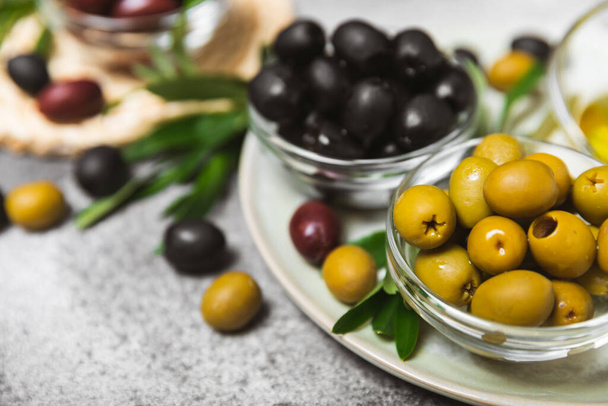 Vihreitä, punaisia ja mustia oliiveja mustalla marmoritaustalla. Erilaisia oliiveja kulhoissa ja oliiviöljyä tuoreita oliivinlehtiä. Herkku.Välimeren keittiö. Kopioi tila. - Valokuva, kuva