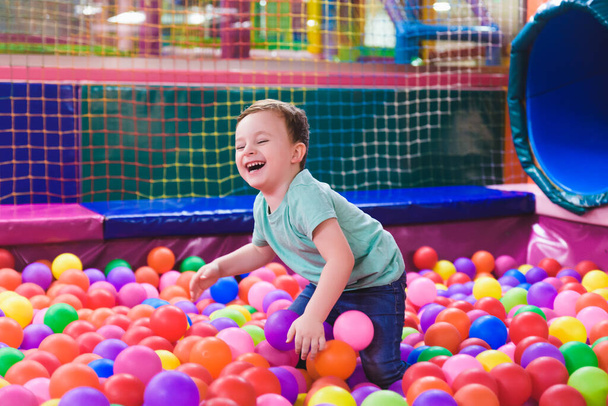 Счастливый смеющийся ребенок смеется в закрытом игровом центре. Дети играют с цветными мячами в бассейне. Партия. - Фото, изображение