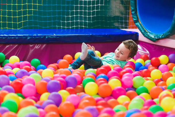 Χαρούμενο γελαστό παιδί που γελάει σε ένα κλειστό κέντρο παιχνιδιού. Τα παιδιά παίζουν με χρωματιστές μπάλες στην πισίνα παιδική χαρά μπάλα. Κόμμα. - Φωτογραφία, εικόνα