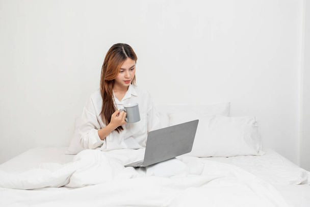 Woman in white nightgown waking up on weekend morning resting and relaxing playing with laptop mobile phone Eating bread and drinking tea in glass inside white bedroom.  Morning vacation concept. - Photo, image