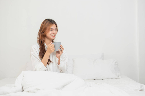 Woman in white nightgown waking up on weekend morning resting and relaxing playing with laptop mobile phone Eating bread and drinking tea in glass inside white bedroom.  Morning vacation concept. - Фото, зображення