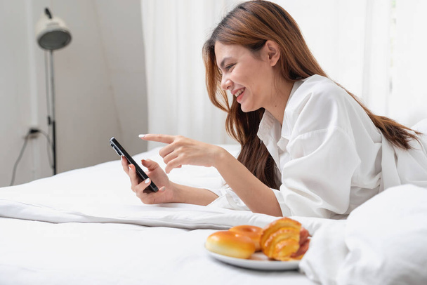 Beyaz gecelikli bir kadın hafta sonu uyanıp dinleniyor ve cep telefonuyla oynuyor ekmek yiyor ve beyaz yatak odasında çay içiyor. Sabah tatili. - Fotoğraf, Görsel