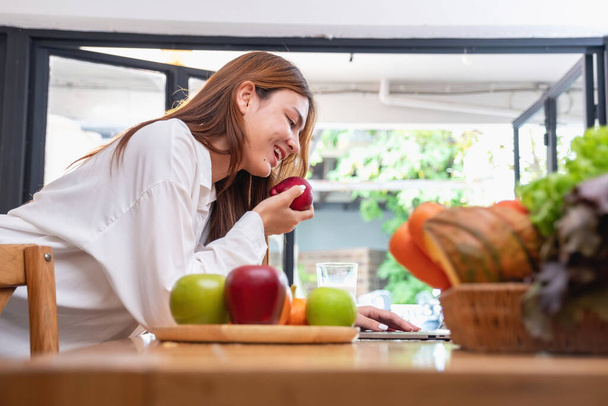 Mujer con una hermosa cara en una camisa blanca está haciendo un desayuno saludable con pan, verduras, frutas y leche dentro de la cocina y la apertura de su computadora portátil para las clases de cocina. ideas de cocina saludable. - Foto, imagen