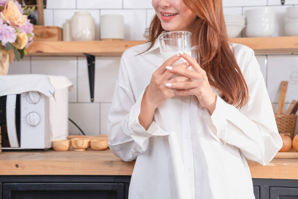 Woman with a beautiful face in a white shirt is making a healthy breakfast with bread, vegetables, fruit and milk inside the kitchen and opening her laptop for cooking lessons. healthy cooking ideas. - Foto, imagen