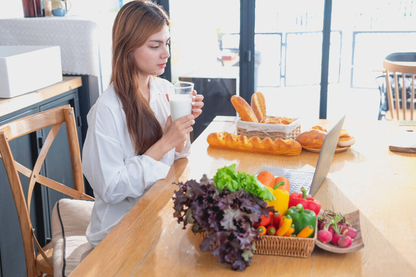Frau mit schönem Gesicht im weißen Hemd bereitet in der Küche ein gesundes Frühstück mit Brot, Gemüse, Obst und Milch zu und öffnet ihren Laptop für Kochkurse. Gesunde Kochideen. - Foto, Bild