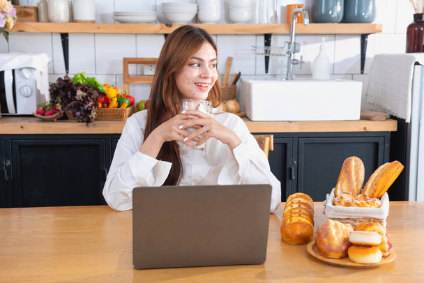 Woman with a beautiful face in a white shirt is making a healthy breakfast with bread, vegetables, fruit and milk inside the kitchen and opening her laptop for cooking lessons. healthy cooking ideas. - 写真・画像
