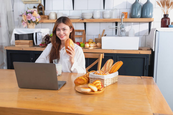Woman with a beautiful face in a white shirt is making a healthy breakfast with bread, vegetables, fruit and milk inside the kitchen and opening her laptop for cooking lessons. healthy cooking ideas. - Photo, Image