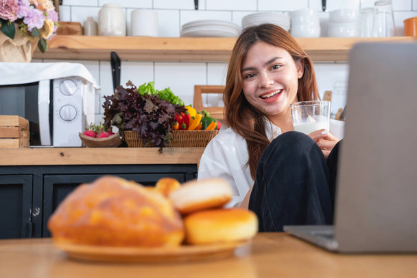 Beyaz gömlekli güzel yüzlü bir kadın mutfakta ekmek, sebze, meyve ve sütle sağlıklı bir kahvaltı hazırlıyor ve yemek dersleri için dizüstü bilgisayarını açıyor. sağlıklı yemek fikirleri. - Fotoğraf, Görsel