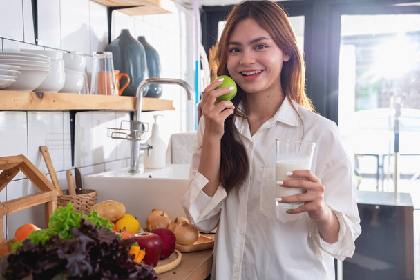 Woman with a beautiful face in a white shirt is making a healthy breakfast with bread, vegetables, fruit and milk inside the kitchen and opening her laptop for cooking lessons. healthy cooking ideas. - Valokuva, kuva