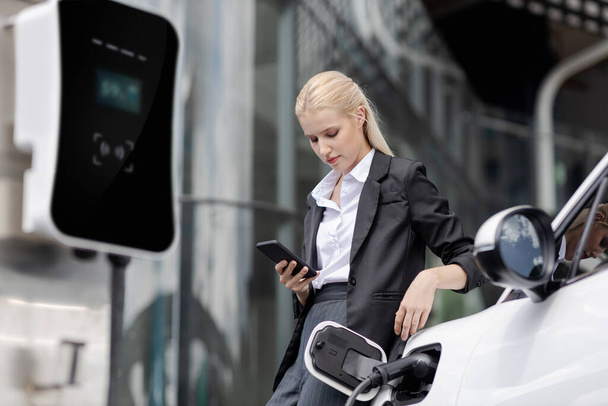 Businesswoman wearing black suit using smartphone, leaning on electric car recharge battery at charging station in city residential building with condos and apartment. Progressive lifestyle concept. - Foto, imagen