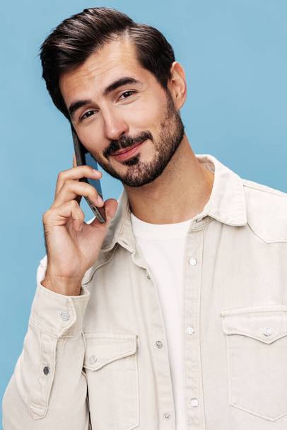 Portrait d'un homme brune gros plan parlant au téléphone sourire avec des dents joie regarde la caméra, sur un fond bleu dans un T-shirt blanc et un jean, espace de copie. Photo de haute qualité - Photo, image