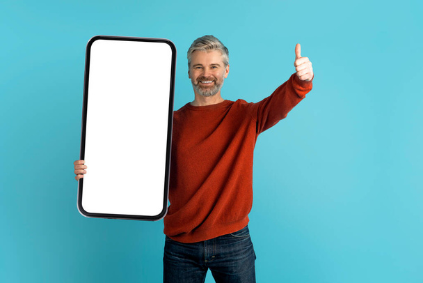 Great offer, nice mobile app. Cheerful handsome grey-haired bearded mature man in casual holding big phone with white blank screen, mockup, showing thumb up, blue studio background - Zdjęcie, obraz