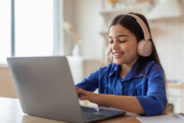 E-Learning Class. Cheerful Arabic Schoolgirl Studying Online Typing On Laptop Computer Wearing Earphones Sitting At Home. School Kid Girl Websurfing Enjoying Distance Lesson - 写真・画像