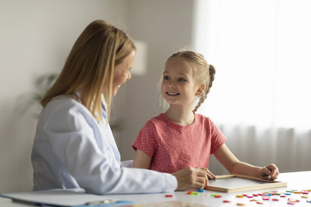 Cute Little Girl Looking At Therapist Lady During Session Meeting In Office, Smiling Female Child Sitting At Desk And Painting On Mini Chalkboard, Enjoying Development Games And Activities, Closeup - Photo, Image