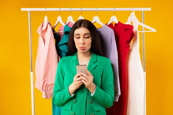 Upset young stylish lady using cellphone while standing near garment rack with colorful clothes, woman upset not to have fashionable outfit, posing on yellow background - Foto, afbeelding