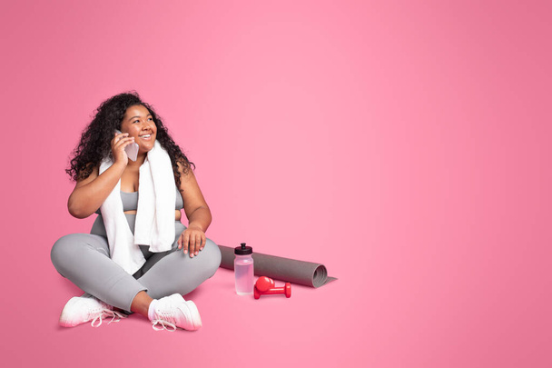 Überdimensionale schwarze Frau in Sportbekleidung, die mit dem Handy telefoniert, mit Hantel und Wasserflasche auf rosa Hintergrund sitzend, freier Raum. Workout-Sportkonzept - Foto, Bild
