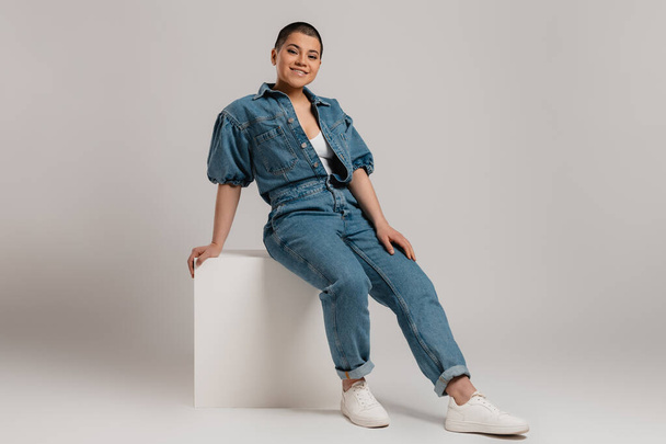 Happy young short hair woman in denim clothes sitting against grey background - Foto, Imagen