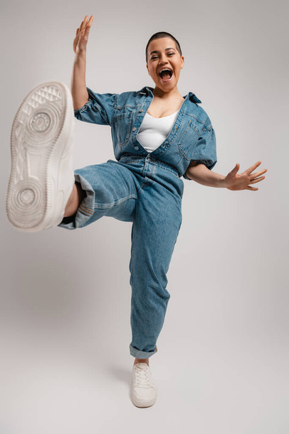 Full length of happy young short hair woman in denim clothes dancing against grey background - Fotoğraf, Görsel