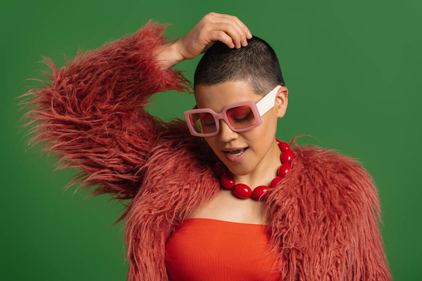 Fashionable young woman in trendy clothes touching her shaved head against green background - Foto, immagini