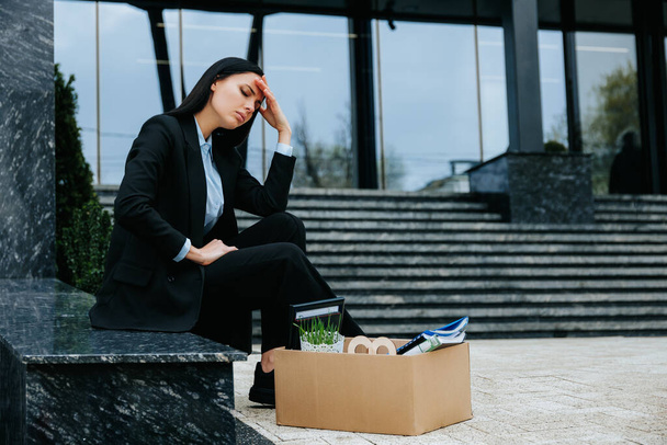 An individual sits on a cardboard box in an outdoor setting, looking unhappy and upset after being fired from their job. Sitting alone outdoors after her dismissal, she reflects on her loss. - Фото, изображение