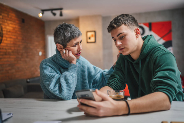 teenage boy and mature caucasian woman sit together at the kitchen at home talk mother and son or relatives support solving problem share experience and opinion boy hold mobile phone smartphone - Foto, imagen