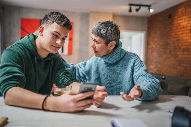 teenage boy and mature caucasian woman sit together at the kitchen at home talk mother and son or relatives support solving problem share experience and opinion boy hold mobile phone smartphone - Foto, Imagen