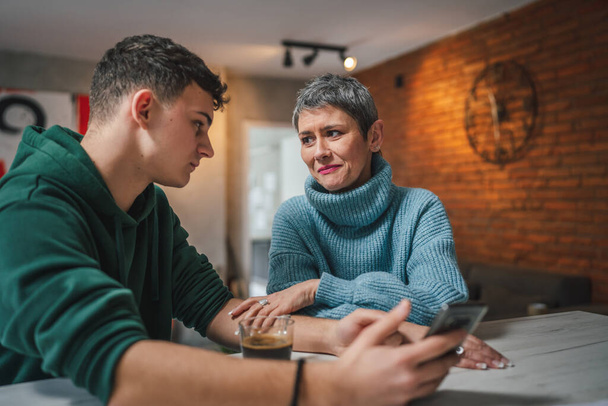 teenage boy and mature caucasian woman sit together at the kitchen at home talk mother and son or relatives support solving problem share experience and opinion boy hold mobile phone smartphone - Foto, afbeelding