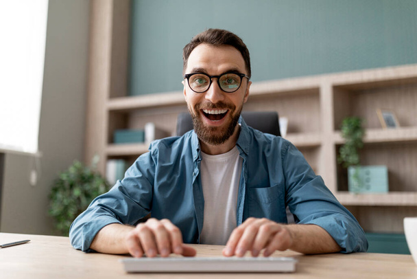 В офисе веселый мужчина-фрилансер смотрит в очки на экране и клавиатуре, реагирует на онлайн-предложение, смотрит на веб-камеру - Фото, изображение