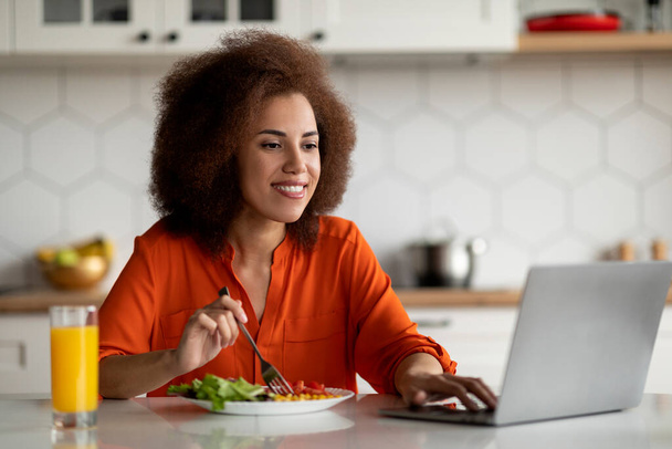 Посмішка молода чорна жінка використовує клапан під час сніданку на кухні, щаслива афро-американська жінка працює віддалено або браузер Інтернет на комп'ютері під час споживання смачної їжі вдома, Копія простору - Фото, зображення