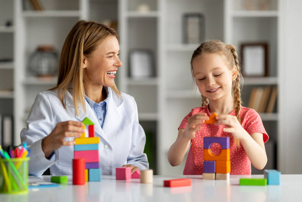 Friendly child development specialist woman having session with cheerful little girl, cute female kid sitting at table and playing with colorful wooden bricks, enjoying learning through play - Photo, Image