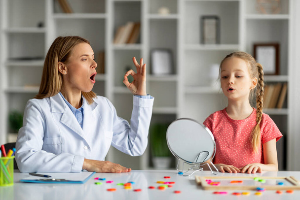 Speech Therapy Concept. Cute little girl pronouncing sound O looking at mirror, professional therapist lady teaching female child right pronounciation during private lesson at office - Fotografie, Obrázek