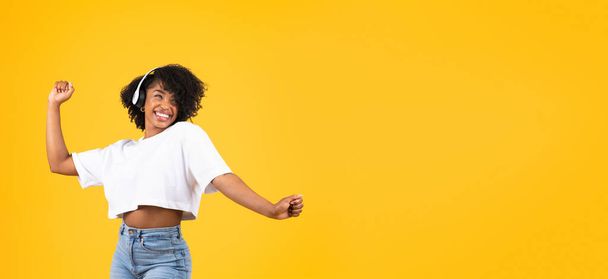 Glad young african american curly woman in wireless headphones dancing, has fun, isolated on yellow studio background, panorama. Leisure time, lady enjoy music and audio app - Photo, Image