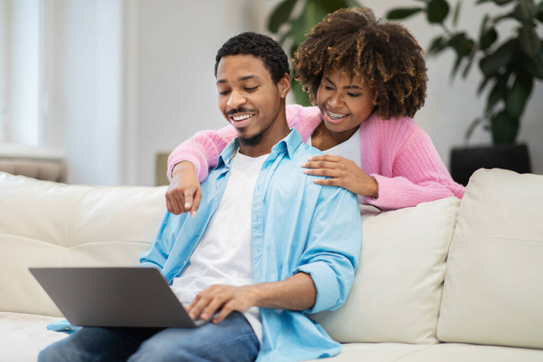 Cheerful young black spouses sitting on couch, planning vacation, using laptop at home. Pretty african american lady hugging her husband from behind and pointing at computer screen, copy space - Фото, изображение